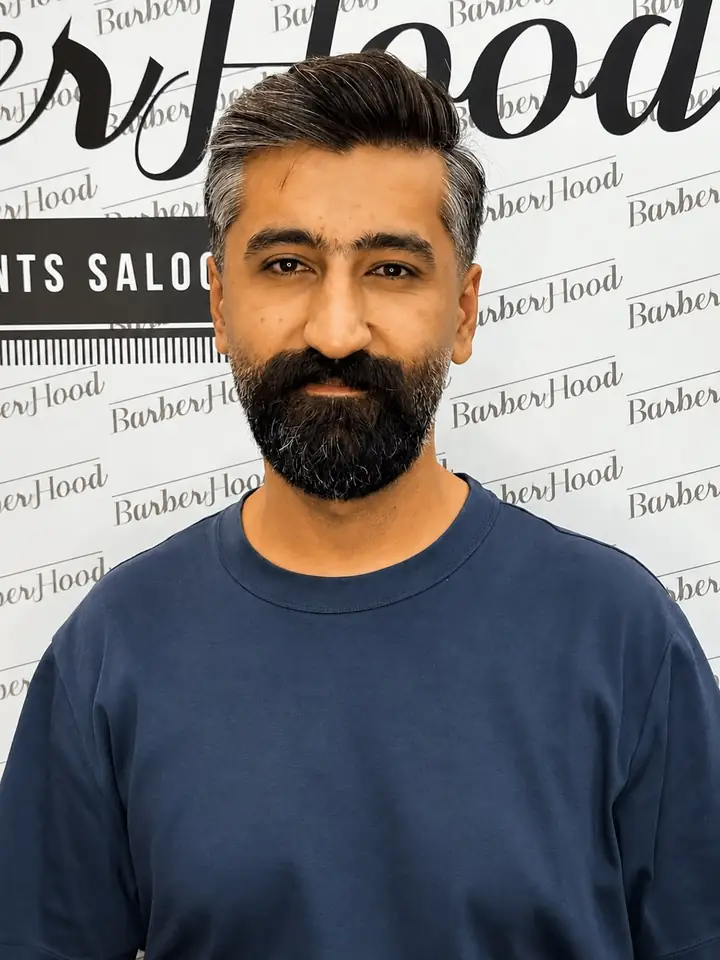 Mohammad, manager at Barberhood in JVC, Dubai — dark hair swept back with grey at the temples and a full beard, wearing a navy t-shirt in front of the branded studio wall